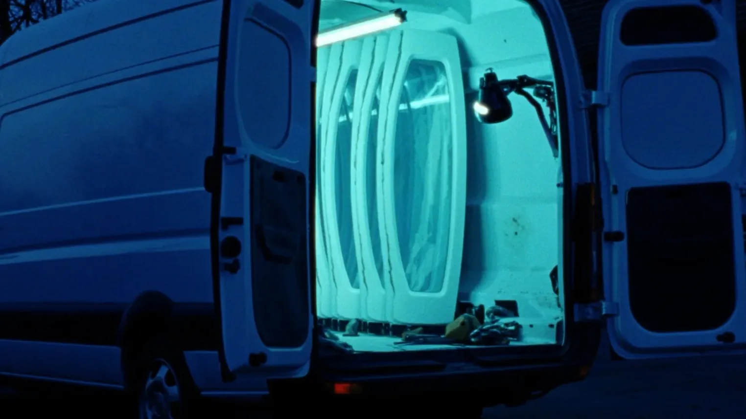The rear doors of the Shalom Auto Glass mobile van open at the curb with a lit rack of windshields inside